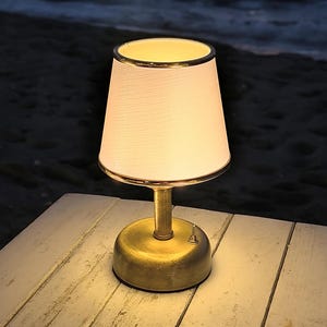 May include: A small, battery-powered table lamp with a white lampshade and a gold-colored base. The lampshade has a gold trim. The lamp is sitting on a white wooden table, with the ocean in the background. The lamp has an on/off switch.