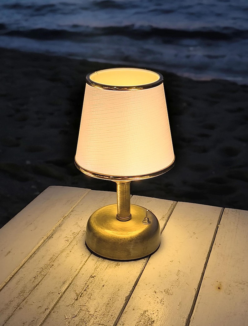 May include: A small, battery-powered table lamp with a white lampshade and a gold-colored base. The lampshade has a gold trim. The lamp is sitting on a white wooden table, with the ocean in the background. The lamp has an on/off switch.