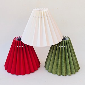 Pode incluir: Três abajures plissados em vermelho, branco e verde. Os abajures têm formato cônico com design plissado. O abajur branco está centralizado, com os abajures vermelho e verde em cada lado. Os abajures são feitos de um material texturizado.