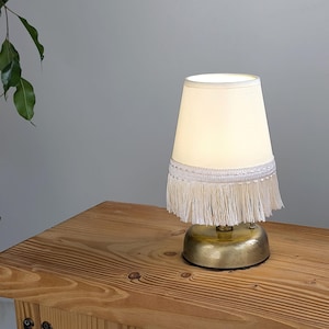 May include: A small, cream-colored table lamp with a white lampshade. The lampshade has a white fringe and sits atop a gold-colored base. The lamp is placed on a wooden table.