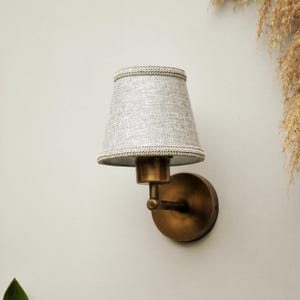 May include: A wall-mounted lamp featuring a gray fabric lampshade and a brass-toned metal base. The shade has a textured finish and is edged with a decorative trim. This lamp is designed for interior lighting and offers a vintage aesthetic.