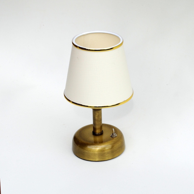 May include: A small, brass-colored table lamp with a cream-colored lampshade. The lampshade has a gold trim and a classic, tapered shape. The lamp has a round base and a small on/off switch. The lamp is designed for indoor use.