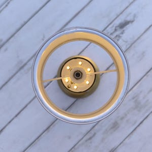 May include: Overhead view of a modern, circular light fixture. The fixture has a clear, cylindrical outer ring with a gold-colored inner ring. Small, warm-toned LED lights are visible at the center, creating a soft glow. The background is a white, wooden surface.