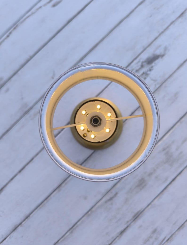 May include: Overhead view of a modern, circular light fixture. The fixture has a clear, cylindrical outer ring with a gold-colored inner ring. Small, warm-toned LED lights are visible at the center, creating a soft glow. The background is a white, wooden surface.
