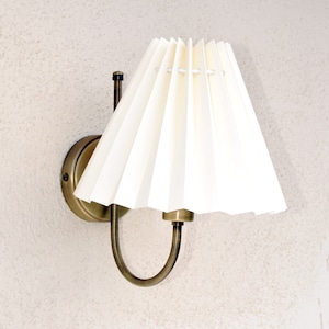 Puede incluir: Una lámpara de pared de latón con una pantalla de tela blanca plisada. La lámpara tiene un diseño sencillo con un brazo curvo y una base redonda.