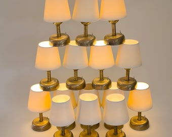 May include: A pyramid arrangement of fifteen small table lamps with brass-toned circular bases and tapered off-white fabric shades. Each lamp features a central stem and a small toggle switch on the base, emitting a warm glow from the light source.