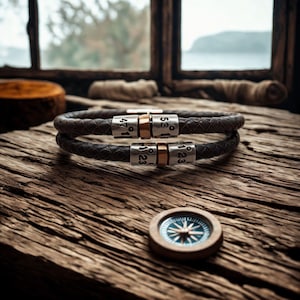 May include: Two dark brown braided leather bracelets with silver and gold details, each with engraved numbers. A small compass with a blue and white design is below the bracelets on a wooden surface. The bracelets are a dark brown colour.