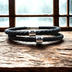 May include: Two black braided leather bracelets with silver metal details. Each bracelet features a silver clasp with the Roman numeral III. The bracelets are presented on a wooden surface.