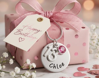 Puede incluir: Un collar con colgante circular de plata grabado con el número 13 y el nombre Chloe, acentuado con un cristal rosa, sobre una caja de regalo rosa con lunares, una cinta rosa y una etiqueta con el texto Happy Birthday y un corazón.