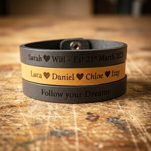 May include: Three stacked leather bracelets with engraved text. The top bracelet is dark grey, the middle is tan, and the bottom is dark grey. The top bracelet reads "Sarah ❤ Will - Est 21st March 2023". The middle bracelet reads "Luca ❤ Daniel ❤ Chloe ❤ Izzy". The bottom bracelet reads "Follow your Dreams".