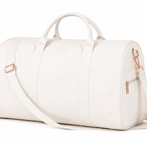 May include: A white duffel bag featuring rose gold hardware, including a side zipper pull, strap buckles, and clips. The bag has two top handles and an adjustable shoulder strap, with a smooth, solid-colored exterior and a structured cylindrical shape.