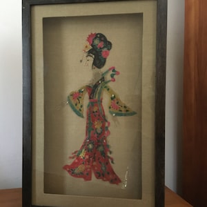 Puede incluir: Obra de arte enmarcada que representa una figura asiática tradicional. La figura lleva un colorido disfraz en capas con detalles florales. La obra está enmarcada en un marco de madera oscura, sobre un fondo neutro.