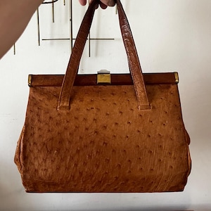 Puede incluir: Un bolso vintage marrón con una superficie texturizada y dos asas marrones. El bolso tiene forma rectangular con un cierre y ribete dorados. El bolso está sostenido por una mano.