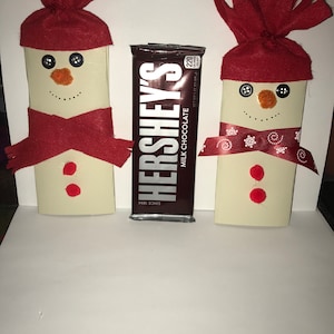 May include: Two handmade snowman decorations with red hats and scarves, featuring button eyes, carrot noses, and painted smiles. A Hershey's Milk Chocolate bar is placed between them. The snowmen have red dot buttons.