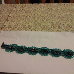 May include: A teal beaded bracelet with flower-shaped designs. Each flower has a dark centre with orange accents, connected by a teal beaded chain. The bracelet has a bronze-coloured clasp. The bracelet is approximately 18 cm in length.