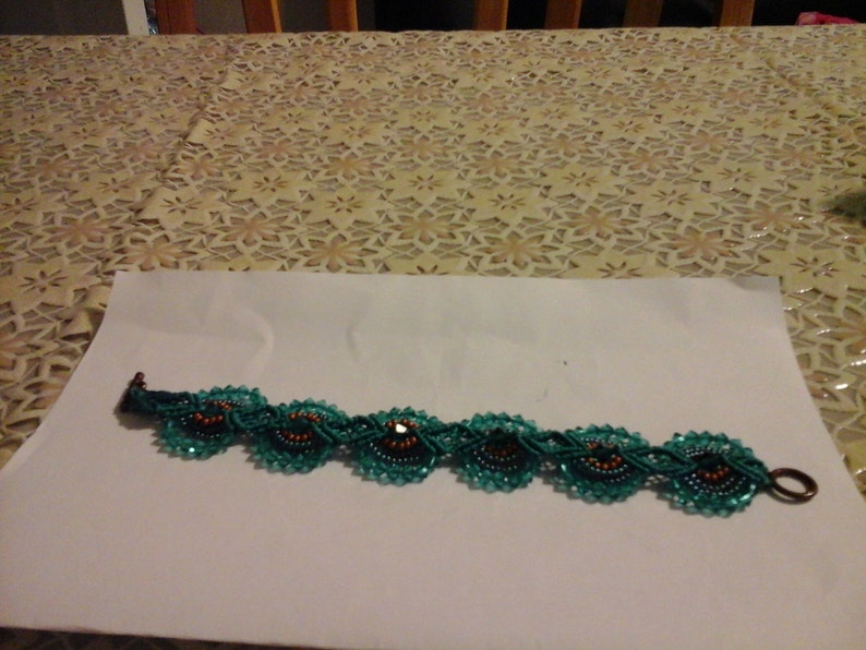 May include: A teal beaded bracelet with flower-shaped designs. Each flower has a dark centre with orange accents, connected by a teal beaded chain. The bracelet has a bronze-coloured clasp. The bracelet is approximately 18 cm in length.