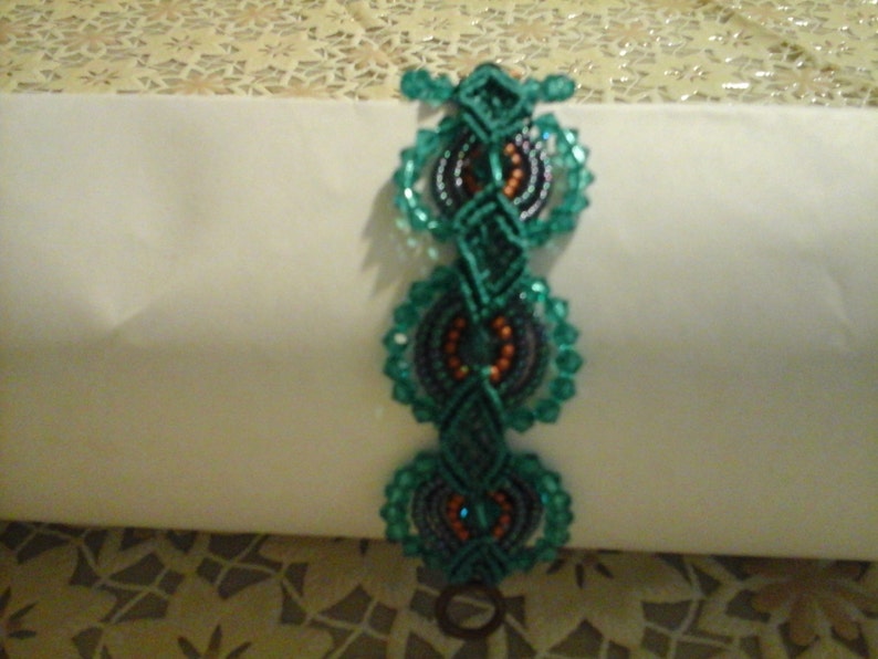 May include: A teal green macrame bracelet, hand-knotted with circular beaded details in teal, orange, and dark blue. The bracelet has a toggle clasp. It is displayed on a white surface.
