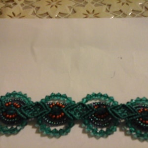 May include: A close-up of a decorative trim featuring a repeating pattern of dark teal macrame circles with orange and black bead accents. The trim is set against a white background with a floral lace border.
