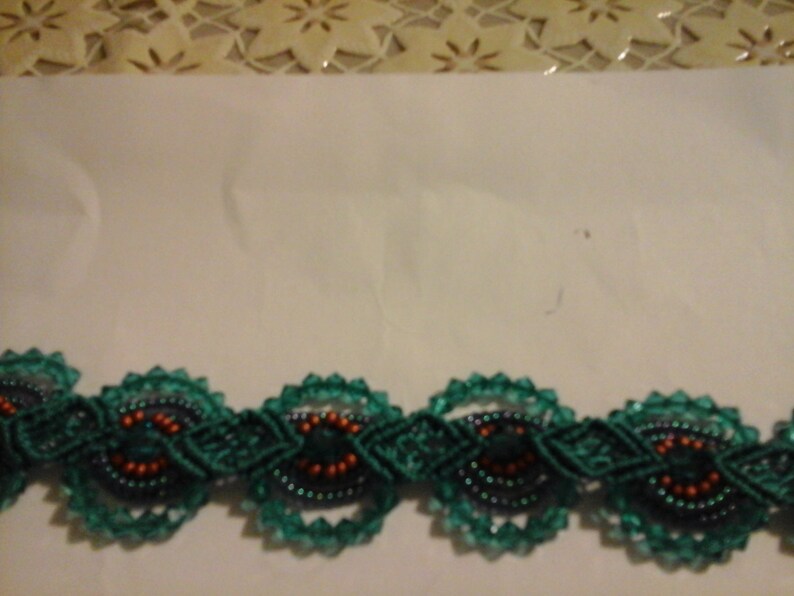 May include: A close-up of a decorative trim featuring a repeating pattern of dark teal macrame circles with orange and black bead accents. The trim is set against a white background with a floral lace border.