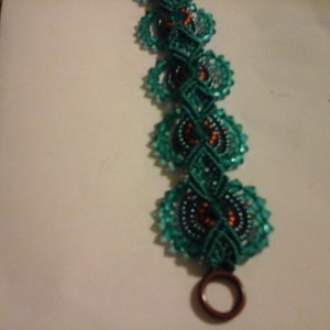 May include: A handmade bracelet with teal circular beaded sections linked by teal macrame. The bracelet has a dark brown clasp. The design incorporates orange and dark blue accents within the circular elements.