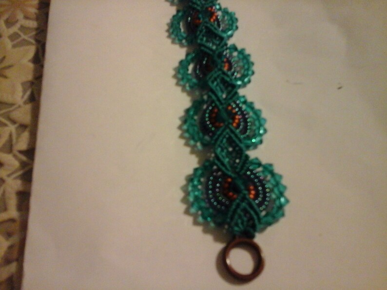 May include: A handmade bracelet with teal circular beaded sections linked by teal macrame. The bracelet has a dark brown clasp. The design incorporates orange and dark blue accents within the circular elements.