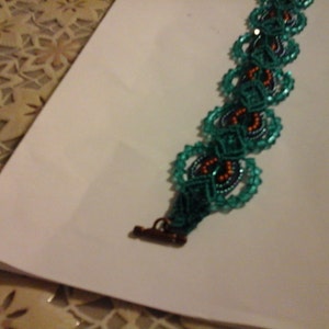 May include: A teal beaded bracelet with a repeating floral design. The bracelet has a bronze clasp and is set against a white background. The centre of each floral shape has orange and dark blue beads. The bracelet is likely around 18 cm in length.