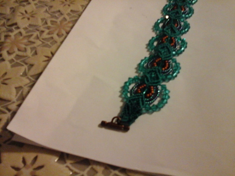 May include: A teal beaded bracelet with a repeating floral design. The bracelet has a bronze clasp and is set against a white background. The centre of each floral shape has orange and dark blue beads. The bracelet is likely around 18 cm in length.