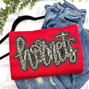 May include: Red t-shirt with a black and leopard print "Mama" design. The shirt is folded and displayed on top of a pair of blue jeans.