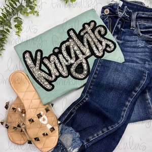 May include: A light blue t-shirt with a silver and black sequined "Knights" applique. The shirt is paired with blue denim jeans and clear sandals with gold studs.