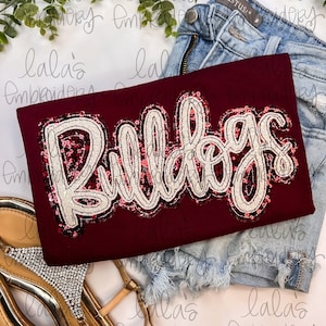 May include: Burgundy t-shirt with a sequined "Bulldogs" design. The shirt is laid on a white surface with a pair of sandals and denim shorts.