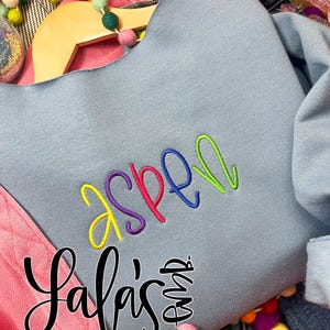 May include: A light blue sweatshirt with the name "Aspen" embroidered in yellow, purple, pink, blue, and green. The sweatshirt is displayed on a wooden hanger. The image also includes the text "Lafa's" in a cursive font.