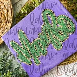 May include: Purple fabric with a green and gold "Mama" applique. The applique is made of a leopard print fabric with green glitter.