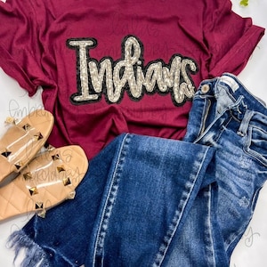 Könnte beinhalten: Ein maroonfarbenes T-Shirt mit einem goldenen und schwarzen Pailletten-Design "Indians". Das Shirt liegt auf einer weißen Oberfläche mit einem Paar transparenter Sandalen und einer blauen Jeans.