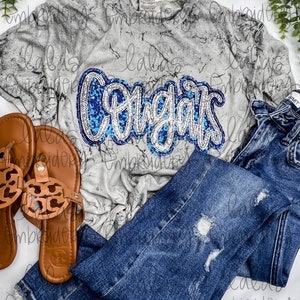 May include: A gray t-shirt with a white marble pattern and a blue and silver sequined "Congrats" design. The shirt is paired with blue jeans.