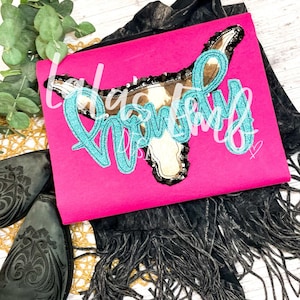 May include: A pink t-shirt with a turquoise glitter design of a cow skull with the word "Howdy" written around it.