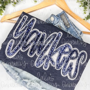 May include: A navy blue t-shirt with a large, sparkly blue and white "Yankees" design. The shirt is hanging on a wooden hanger and is paired with a pair of light blue denim shorts.