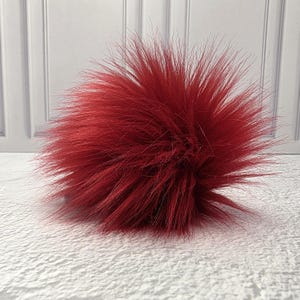 May include: A round, fluffy pom-pom crafted from long, deep red synthetic fibers. The spherical accessory features a dense, textured appearance with individual strands radiating outward from the center, creating a voluminous and soft visual effect.