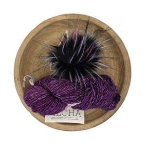 May include: A wooden bowl holds a skein of deep purple yarn, a black and white faux fur pom-pom, and a tag. The yarn is a rich, variegated color. The pom-pom has accents of pink and white. The tag reads "LCHA".