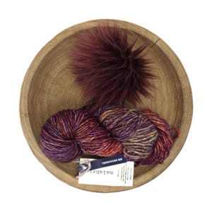 May include: A wooden bowl holds a skein of variegated yarn in shades of purple, red, and gold, along with a fluffy, dark burgundy pompom. A small tag with text is attached to the yarn. The bowl is set against a white background.