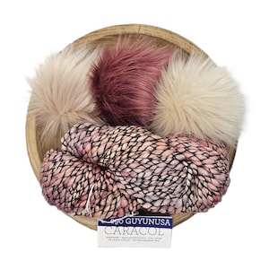 May include: A wooden bowl contains three fluffy pom-poms in shades of pink and cream, along with a skein of pink and cream yarn. The yarn is labeled "CARACOL" and "ego GUYUNUSA". The items are arranged on a white background.
