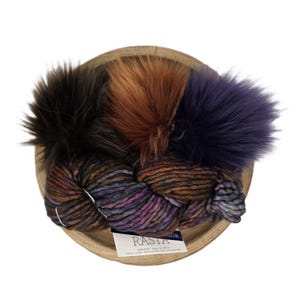 May include: A wooden bowl filled with colorful yarn and three faux fur pom-poms. The yarn is a mix of purple, brown, and orange hues. The pom-poms are black, orange, and dark purple. A label with the word "RASTA" is visible.