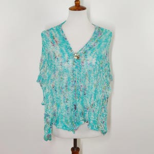 May include: A turquoise, open-weave shawl with a V-neck design. The shawl is fastened with a decorative brooch. The fabric has a speckled pattern with hints of purple and white. The garment is displayed on a mannequin.