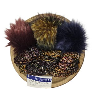 May include: A wooden bowl holds three faux fur pom-poms in burgundy, gold, and navy blue, along with a skein of speckled yarn. The yarn label reads "862 PIEDRAS CARACOL". The yarn is a mix of purple, gold, and brown.