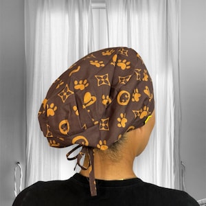 May include: Brown scrub hat with a brown and gold pattern of paw prints and a repeated geometric design. The hat has a tie at the back.