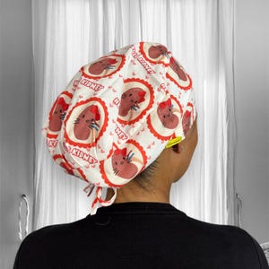 Op de afbeelding: Een witte chirurgische scrub-muts met een rode en bruine print van een cartoonnier met de tekst "I ♥ Kidney" die over het hele ontwerp wordt herhaald.