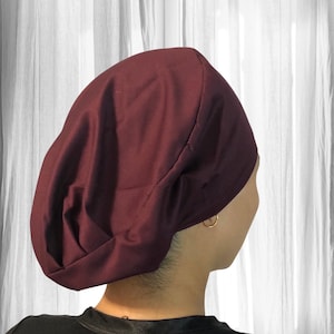 May include: A solid deep burgundy scrub cap featuring a rounded, slouchy silhouette with a wide, structured band around the forehead. The fabric is smooth and opaque, providing full coverage for the head with soft folds at the back.