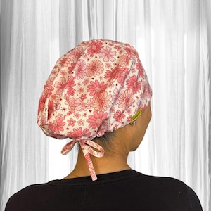 May include: A pink and white floral patterned scrub cap with a tie closure. The cap features a repeating pattern of pink flowers and is worn by a person wearing a black shirt.