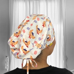 May include: A white scrub cap with a repeating pattern of cartoon croissants, macarons, and mustachioed faces. The cap has a tie closure and is made of a lightweight fabric. The design features orange, red, purple, and blue accents.