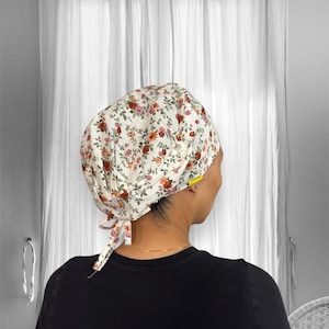 May include: A scrub cap featuring a cream-colored background with a floral pattern of small red, orange, and pink flowers with green leaves. The cap has a rounded crown, a gathered back, and two fabric ties hanging down from the base of the head.