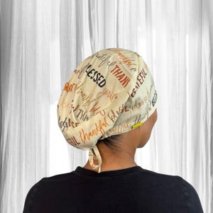 Puede incluir: Gorro de quirófano color crema con las palabras "Blessed", "Thankful" y "Grateful" en escritura marrón y naranja. El gorro está atado en la parte posterior. Se muestra una persona con una camiseta negra.